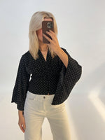 Person tager selfie, iført sort prikket skjorte og hvide bukser, står foran en lys baggrund.