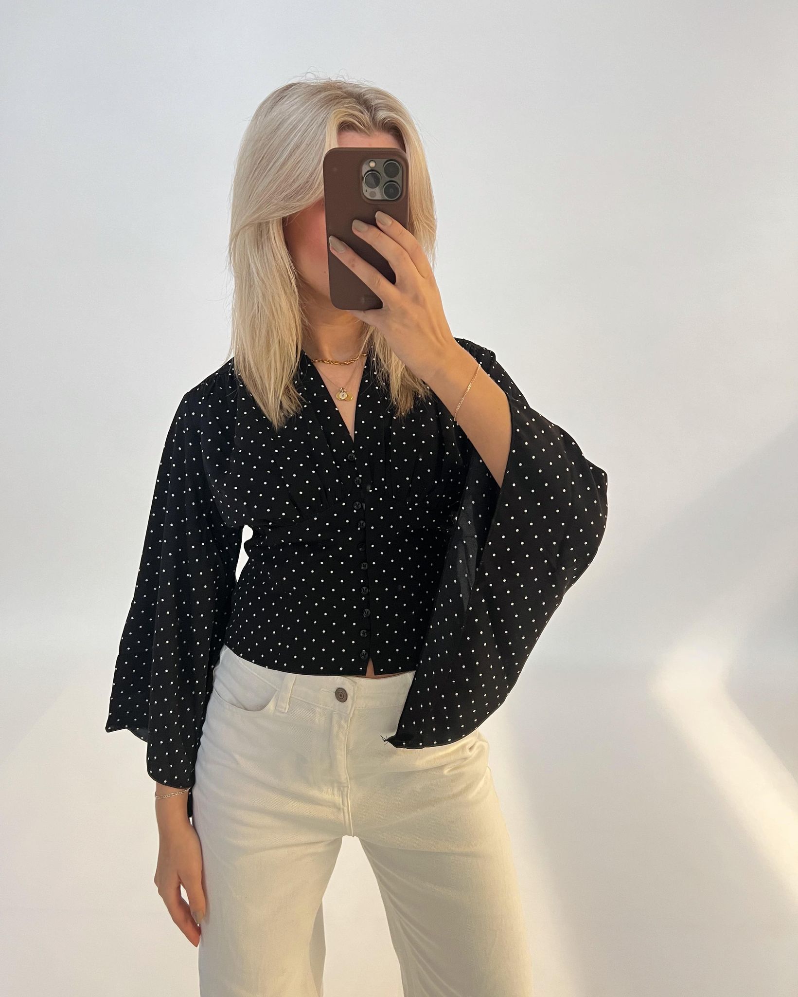 Person tager selfie, iført sort prikket skjorte og hvide bukser, står foran en lys baggrund.