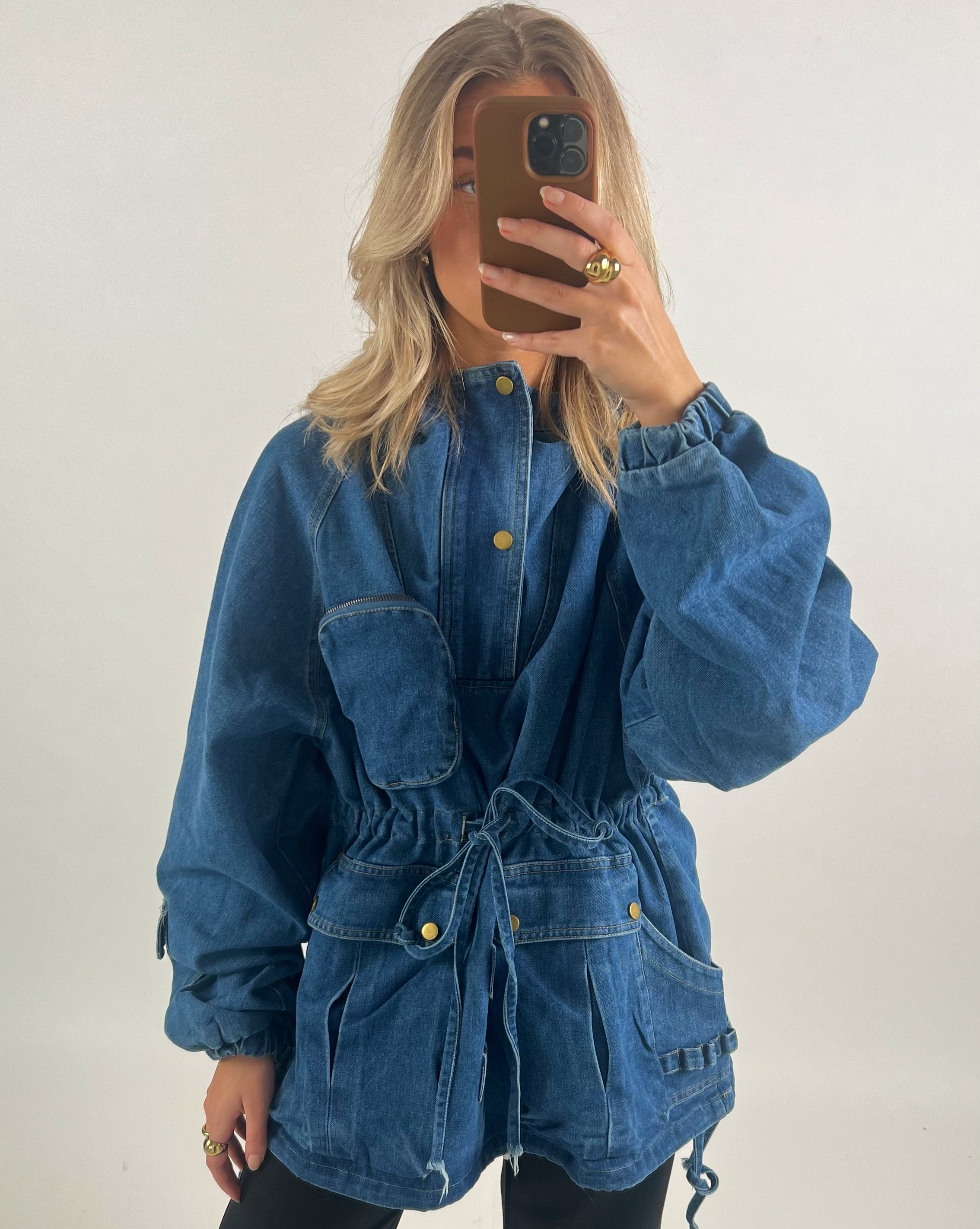 En person iført en stor, blå denimjakke med justerbare snore og lommer tager et spejlbillede med en mobiltelefon i et minimalistisk rum.