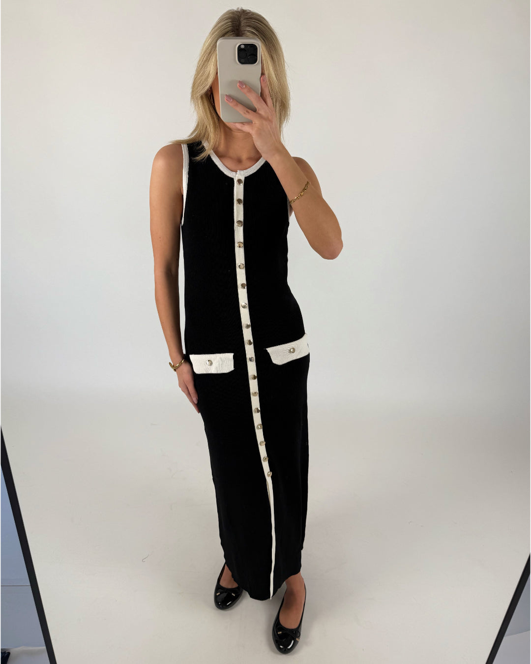 En person iført en lang, sort, ærmeløs kjole med hvide detaljer og knapper står og tager et spejlfoto. Personen er i et rum med en lys, neutral baggrund.