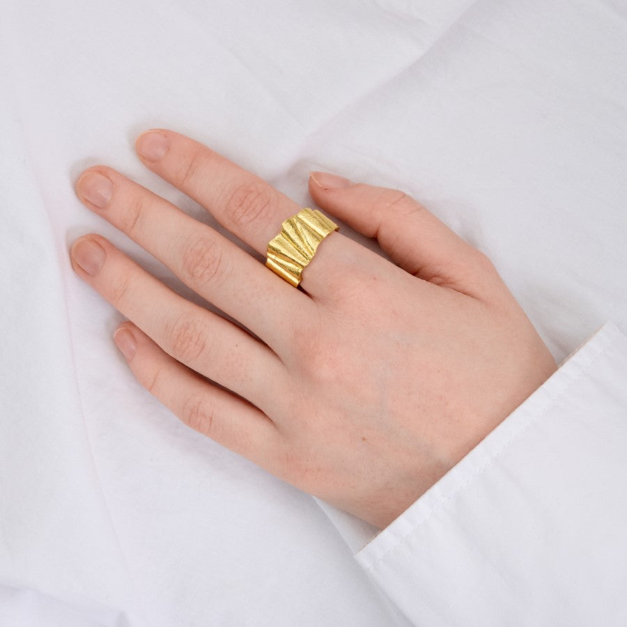 En hånd med en gylden ring med bølget design sidder afslappet på et hvidt klæde, delvist dækket af en hvid skjorteærme.