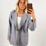 En person iført en lysegrå blazer og hvid top tager en selfie med en brun telefon mod en neutral, lys baggrund.