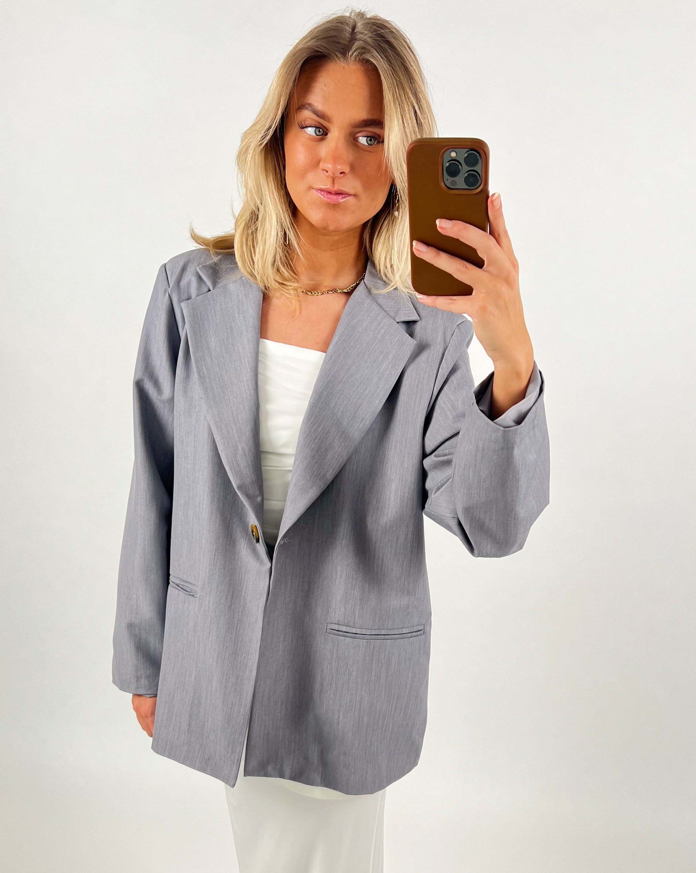 En person iført en lysegrå blazer og hvid top tager en selfie med en brun telefon mod en neutral, lys baggrund.
