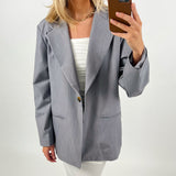 En person iført en grå blazer tager et spejlbillede med en smartphone. Baggrunden er neutral, og personen bærer en hvid top og guldhalskæde.
