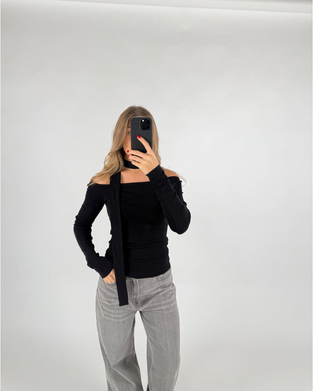 En person iført sort off-shoulder trøje og grå bukser tager et spejlfoto med en smartphone, stående i et minimalistisk, hvidt rum.