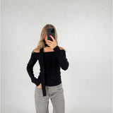 En person iført sort off-shoulder trøje og grå bukser tager et spejlfoto med en smartphone, stående i et minimalistisk, hvidt rum.