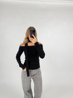 En person iført sort off-shoulder trøje og grå bukser tager et spejlfoto med en smartphone, stående i et minimalistisk, hvidt rum.
