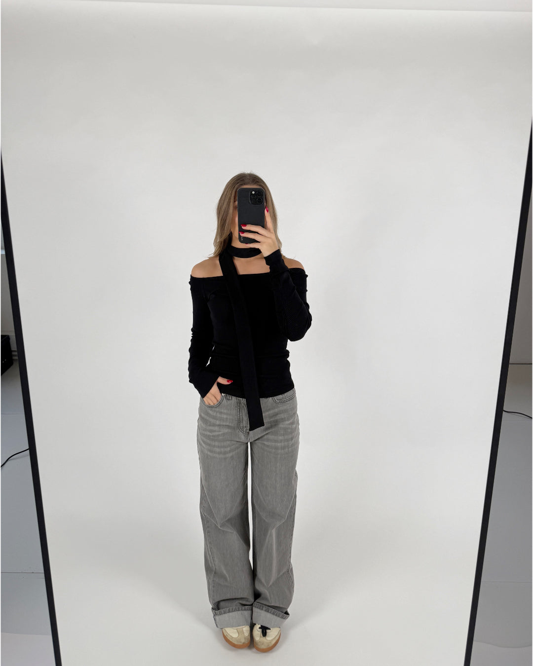 En person tager et spejlbillede, iført en sort off-shoulder top og grå jeans, i et hvidt rum med en enkel baggrund. Personen holder en sort telefon.