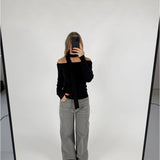 En person tager et spejlbillede, iført en sort off-shoulder top og grå jeans, i et hvidt rum med en enkel baggrund. Personen holder en sort telefon.