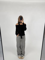 En person tager et spejlbillede, iført en sort off-shoulder top og grå jeans, i et hvidt rum med en enkel baggrund. Personen holder en sort telefon.