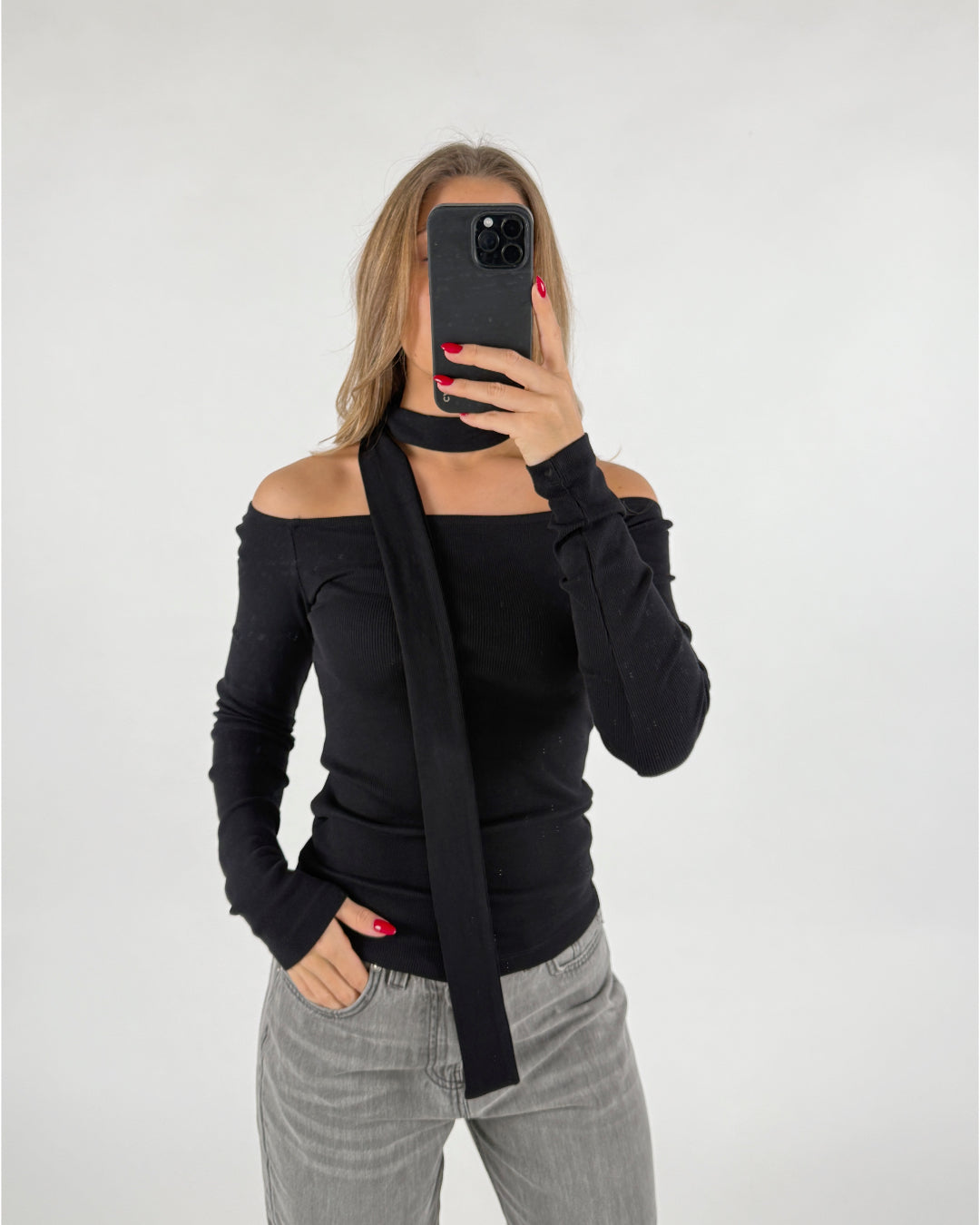 En person står foran et hvidt baggrund iført en sort off-shoulder trøje med et langt halstørklæde, holder en mobiltelefon for at tage et billede, en hånd i bukselommen.