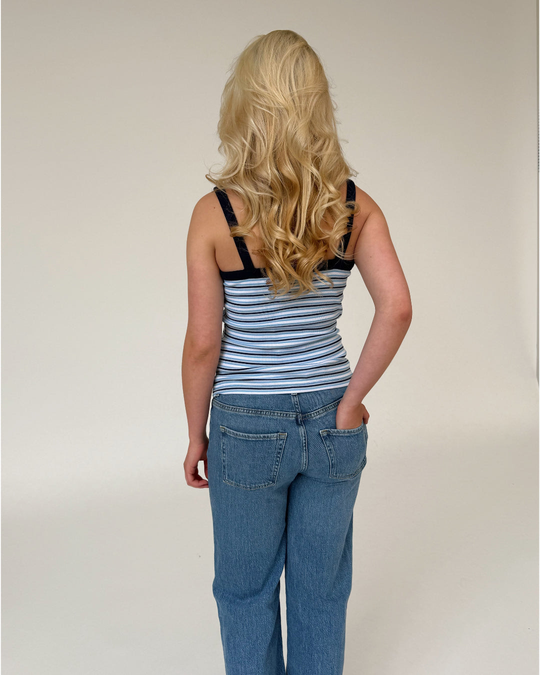 En person med langt, blondt, bølget hår står med ryggen til, iført en stribet tanktop og jeans, på en neutral, lys baggrund.