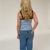 En person med langt, blondt, bølget hår står med ryggen til, iført en stribet tanktop og jeans, på en neutral, lys baggrund.