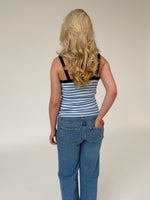 En person med langt, blondt, bølget hår står med ryggen til, iført en stribet tanktop og jeans, på en neutral, lys baggrund.