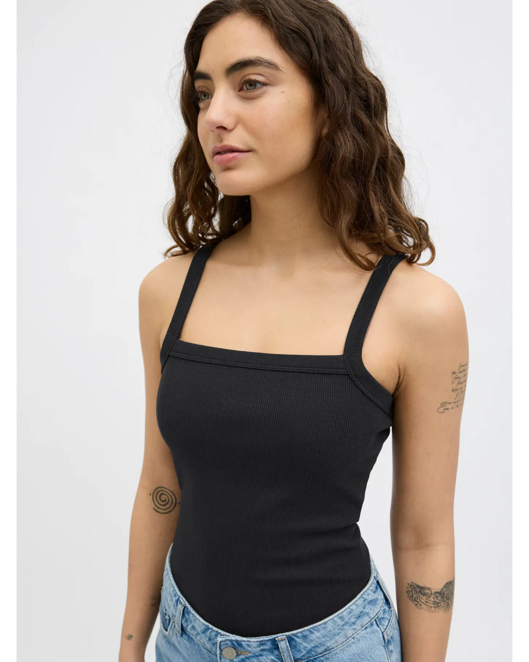Kvinde iført sort tanktop står med afslappet udtryk, omgivet af en neutral grå baggrund. Hun har flere tatoveringer på armen og bærer lyseblå jeans.