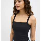 Kvinde iført sort tanktop står med afslappet udtryk, omgivet af en neutral grå baggrund. Hun har flere tatoveringer på armen og bærer lyseblå jeans.