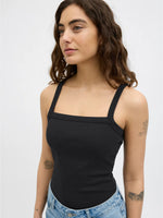 Kvinde iført sort tanktop står med afslappet udtryk, omgivet af en neutral grå baggrund. Hun har flere tatoveringer på armen og bærer lyseblå jeans.