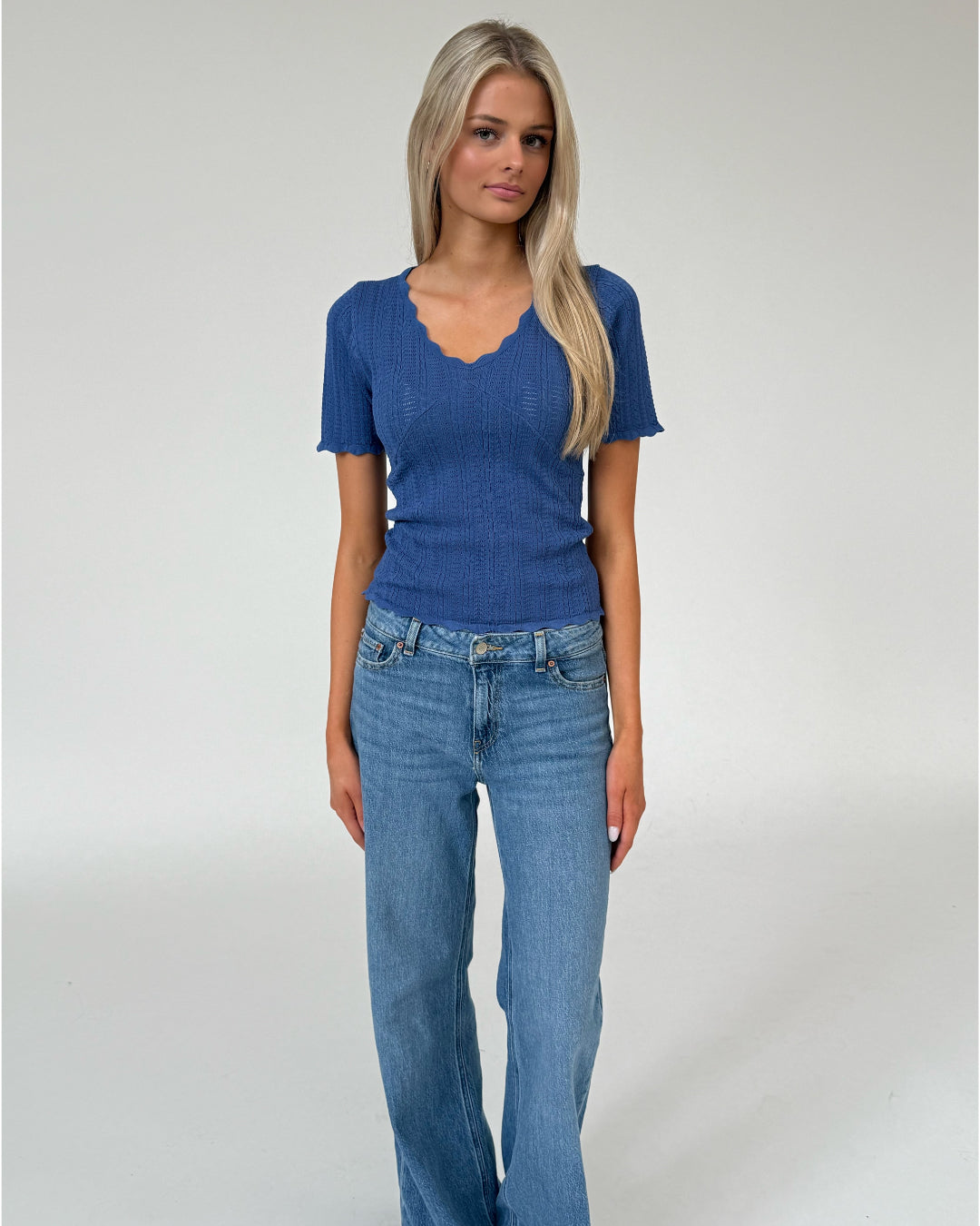 Kvinde står iført en blå, ribbet bluse og lyse jeans. Blusen har bølget kant og v-hals. Baggrunden er en simpel, lys grå væg.