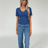 Kvinde står iført en blå, ribbet bluse og lyse jeans. Blusen har bølget kant og v-hals. Baggrunden er en simpel, lys grå væg.