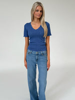 Kvinde står iført en blå, ribbet bluse og lyse jeans. Blusen har bølget kant og v-hals. Baggrunden er en simpel, lys grå væg.