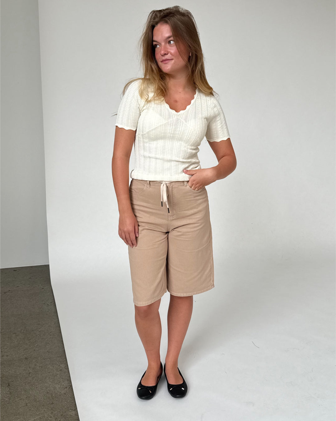 En kvinde står afslappet iført en hvid trøje og beige shorts, med hånden i lommen, på en baggrund af en hvid væg og gråt gulv.