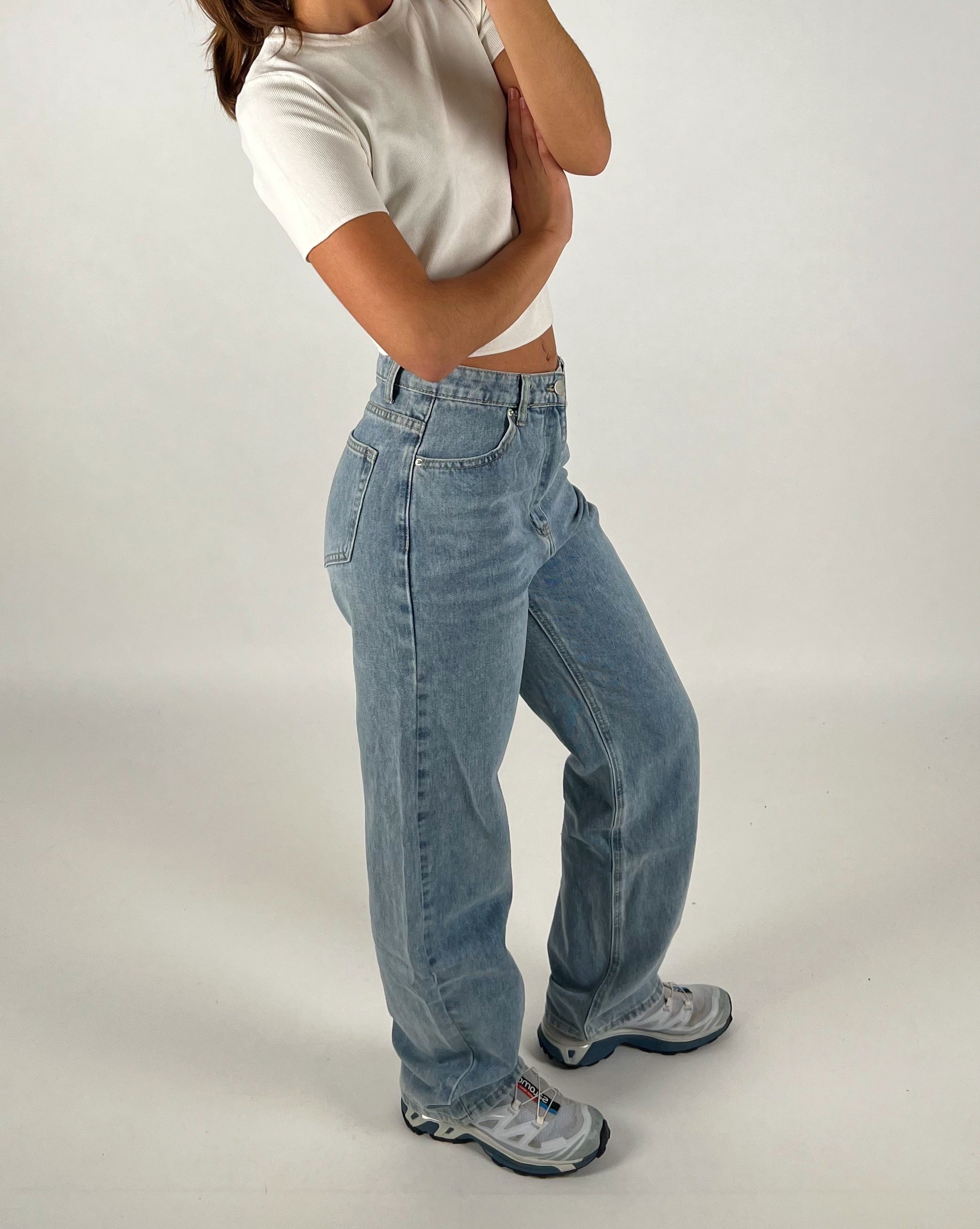 En person iført en hvid t-shirt og lyseblå jeans står i en afslappet stilling. Personen har armene krydset, står på et hvidt baggrund og bærer sportssko.