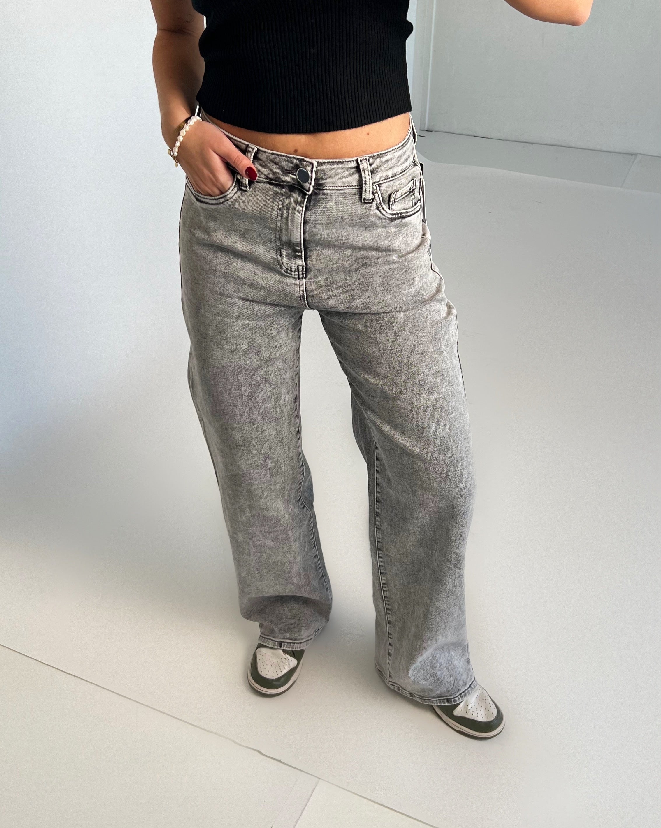 En person iført grå jeans står afslappet med hænderne i lommerne. Personen bærer en sort top og hvide sneakers med sorte detaljer på et lyst gulv.