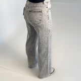 Grå jeans bæres af en person iført sort bluse og grønne sko. Personen står i et lysegråt rum med hvidt gulv.