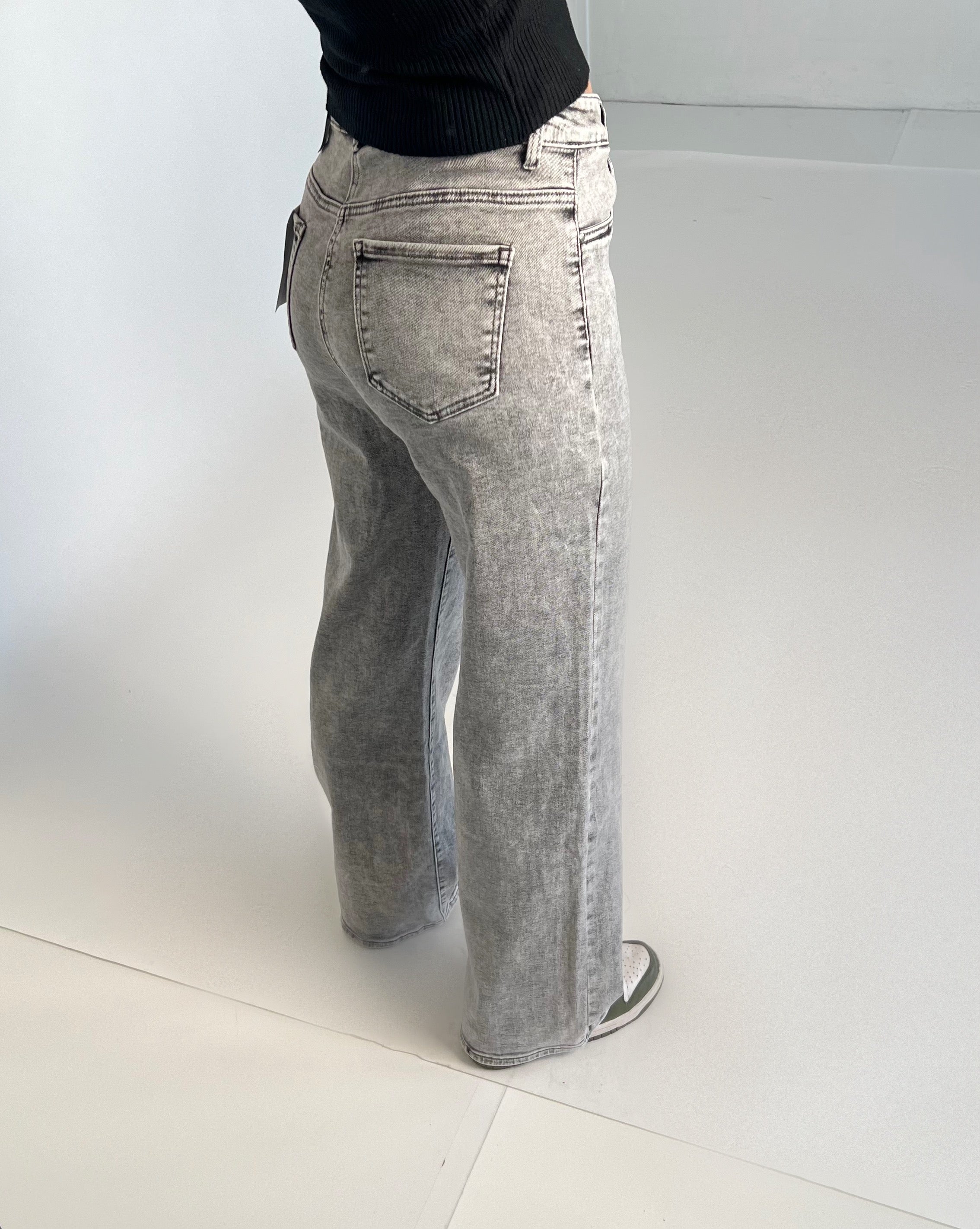 Grå jeans bæres af en person iført sort bluse og grønne sko. Personen står i et lysegråt rum med hvidt gulv.