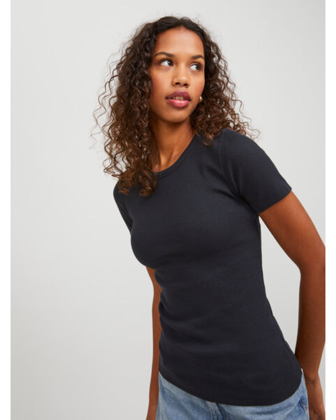 En kvinde med krøllet hår står iført en sort T-shirt og jeans. Hun kigger til siden i et minimalistisk indendørs miljø med en grå baggrund.