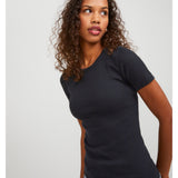 En kvinde med krøllet hår står iført en sort T-shirt og jeans. Hun kigger til siden i et minimalistisk indendørs miljø med en grå baggrund.