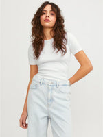 En person står afslappet iført en hvid T-shirt og lyse denim jeans. Konteksten er en simpel, lys baggrund. Ingen tekst er til stede.