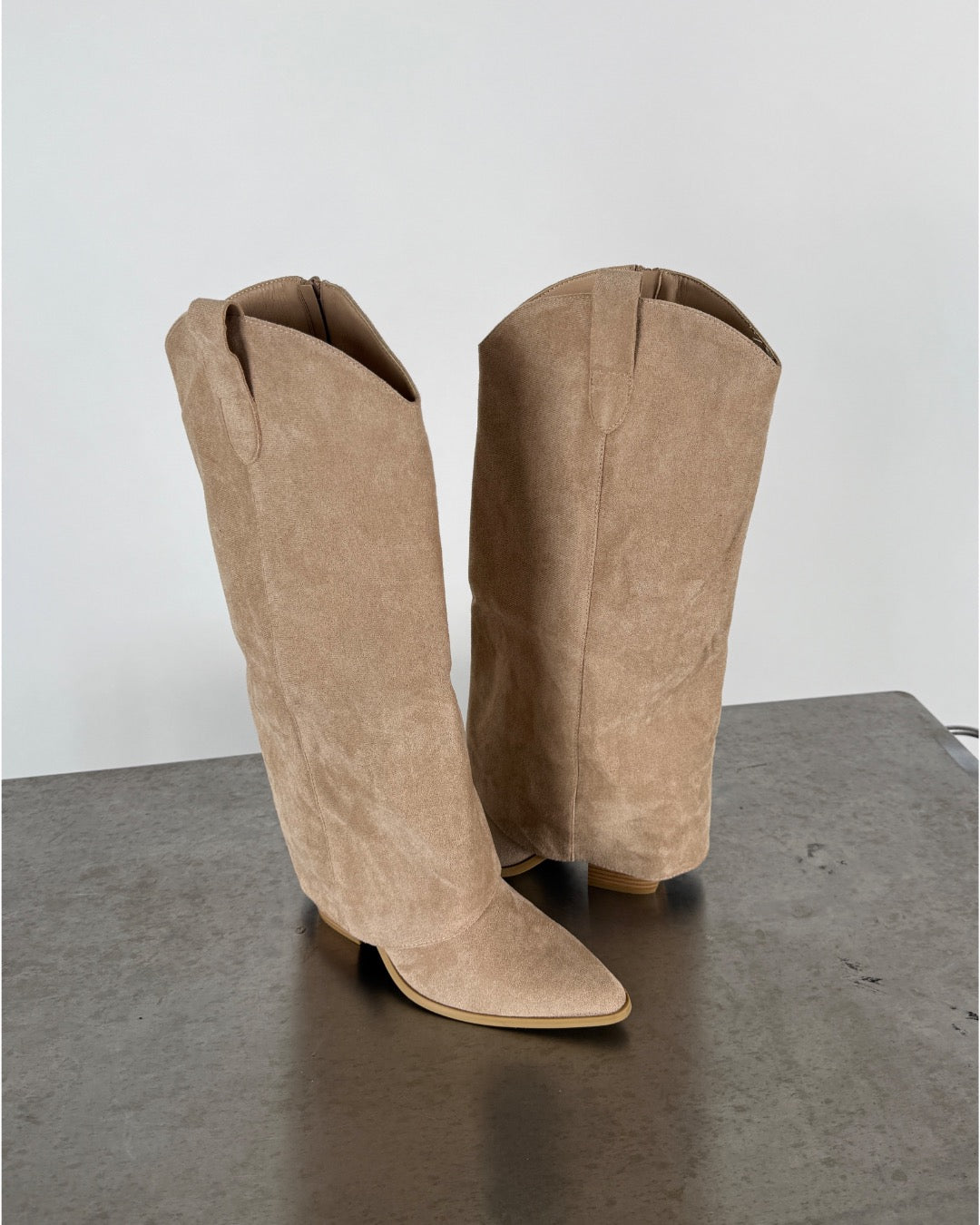 To beige støvler med spidse snuder står oprejst på en grå overflade, i en simpel og minimalistisk indendørs baggrund.