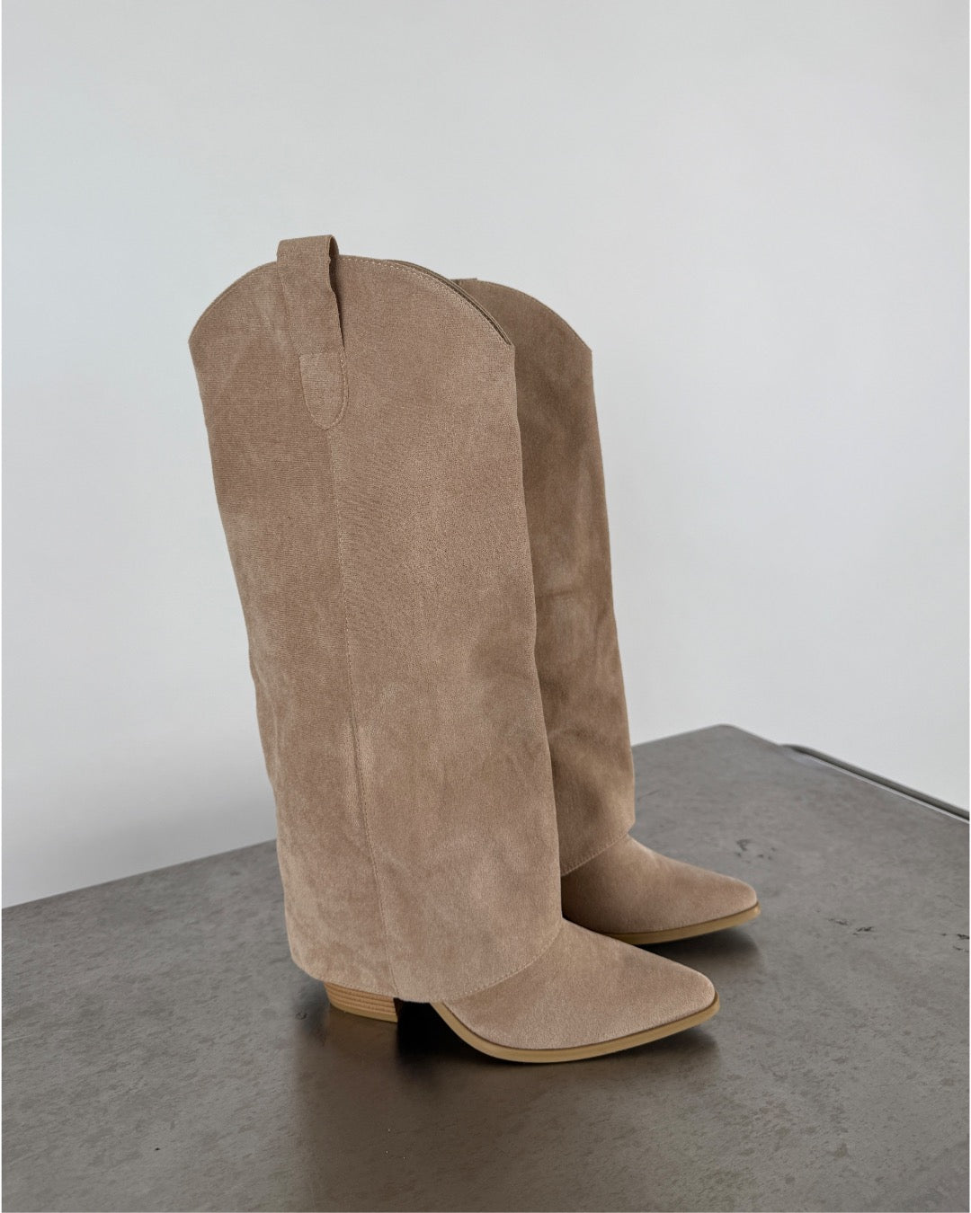 Beige cowboystøvler står på en grå overflade i et neutralt rum, med spidse tæer og lave hæle. Ingen tekst er synlig.