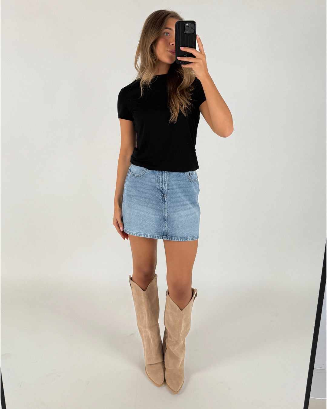 En person iført sort T-shirt, lyseblå denimnederdel og beige støvler tager et spejlbillede i et lyst rum.
