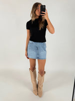 En person iført sort T-shirt, lyseblå denimnederdel og beige støvler tager et spejlbillede i et lyst rum.