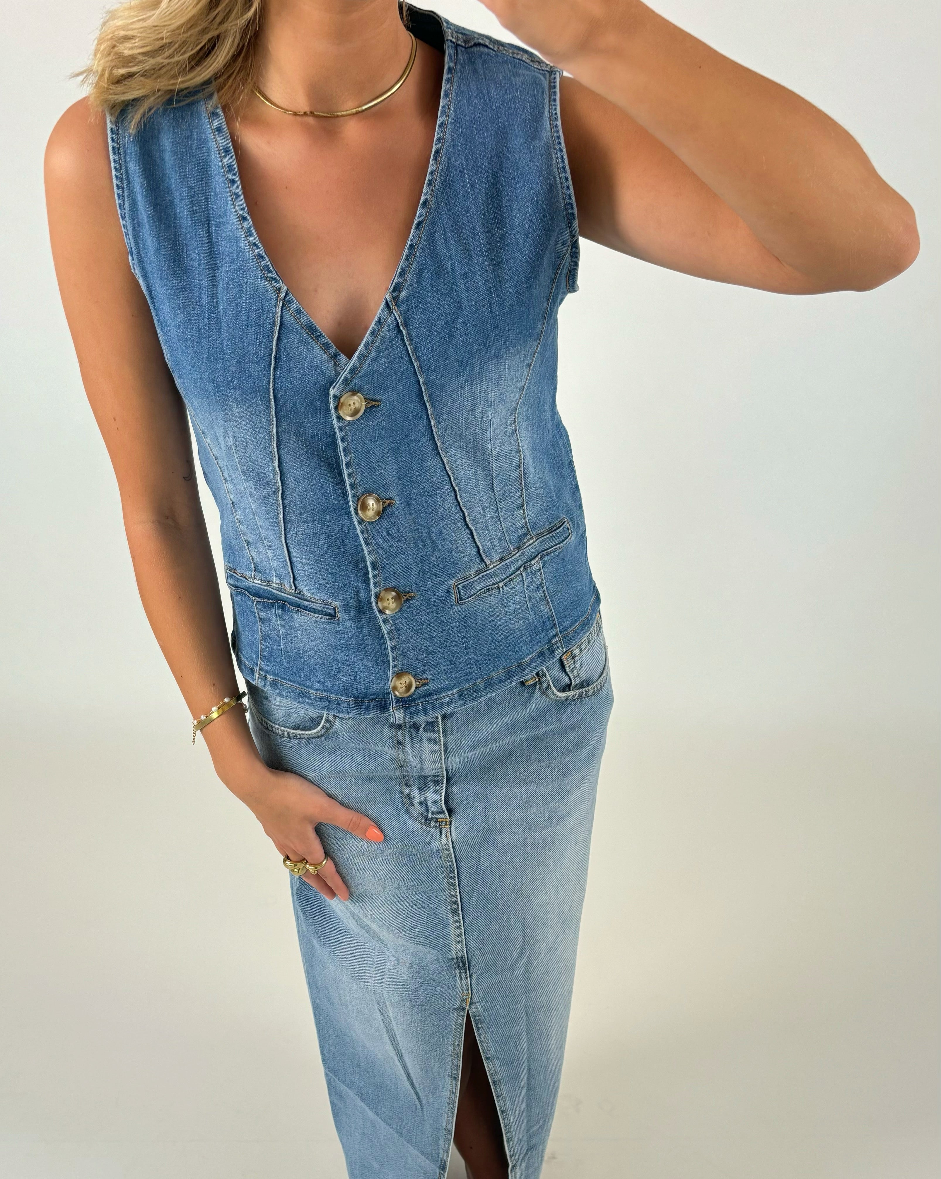 En person iført en ærmeløs denimvest med knapper og en matchende denimnederdel med slids. Personen har et armbånd og en halskæde på, mod en lys baggrund.