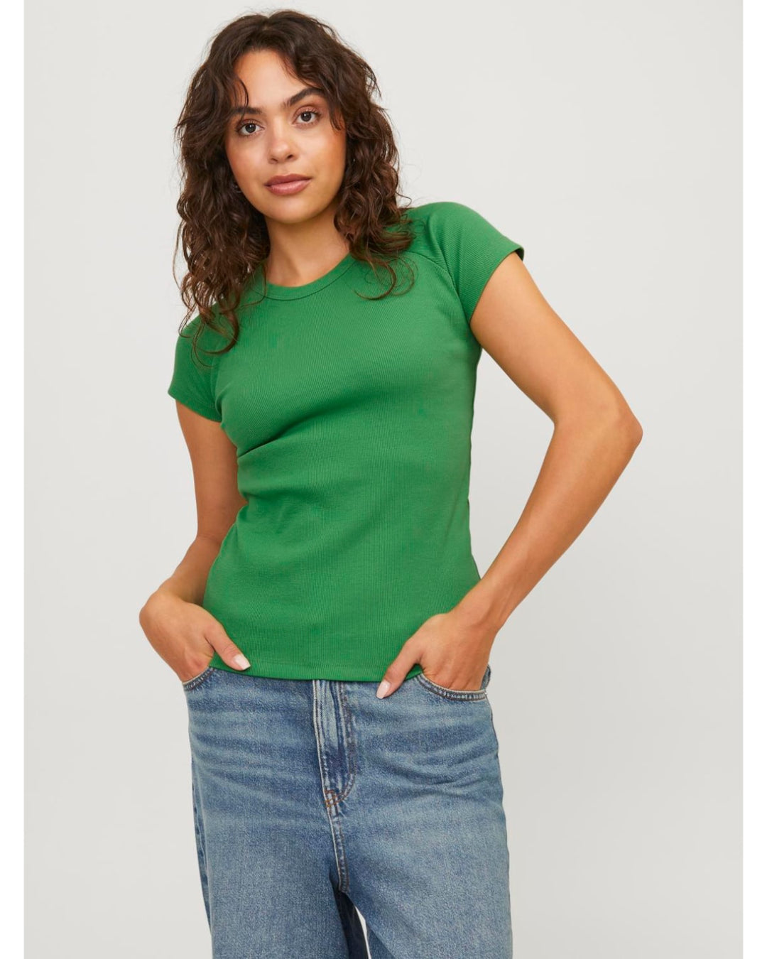 En person står iført en grøn t-shirt og blå jeans, med hænderne i lommerne, mod en lys baggrund. Persoen har krøllet hår og ser direkte fremad.