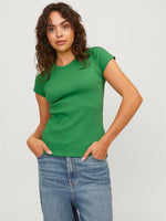 En person står iført en grøn t-shirt og blå jeans, med hænderne i lommerne, mod en lys baggrund. Persoen har krøllet hår og ser direkte fremad.