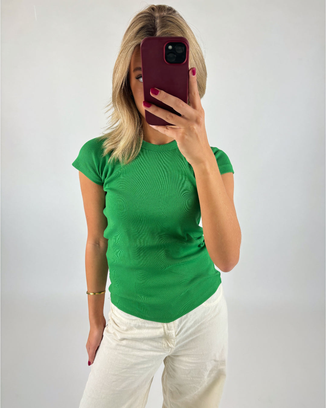 En person iført en grøn t-shirt tager en selfie med en smartphone i rød etui, står mod en neutral væg.