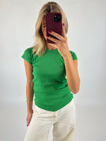 En person iført en grøn t-shirt tager en selfie med en smartphone i rød etui, står mod en neutral væg.