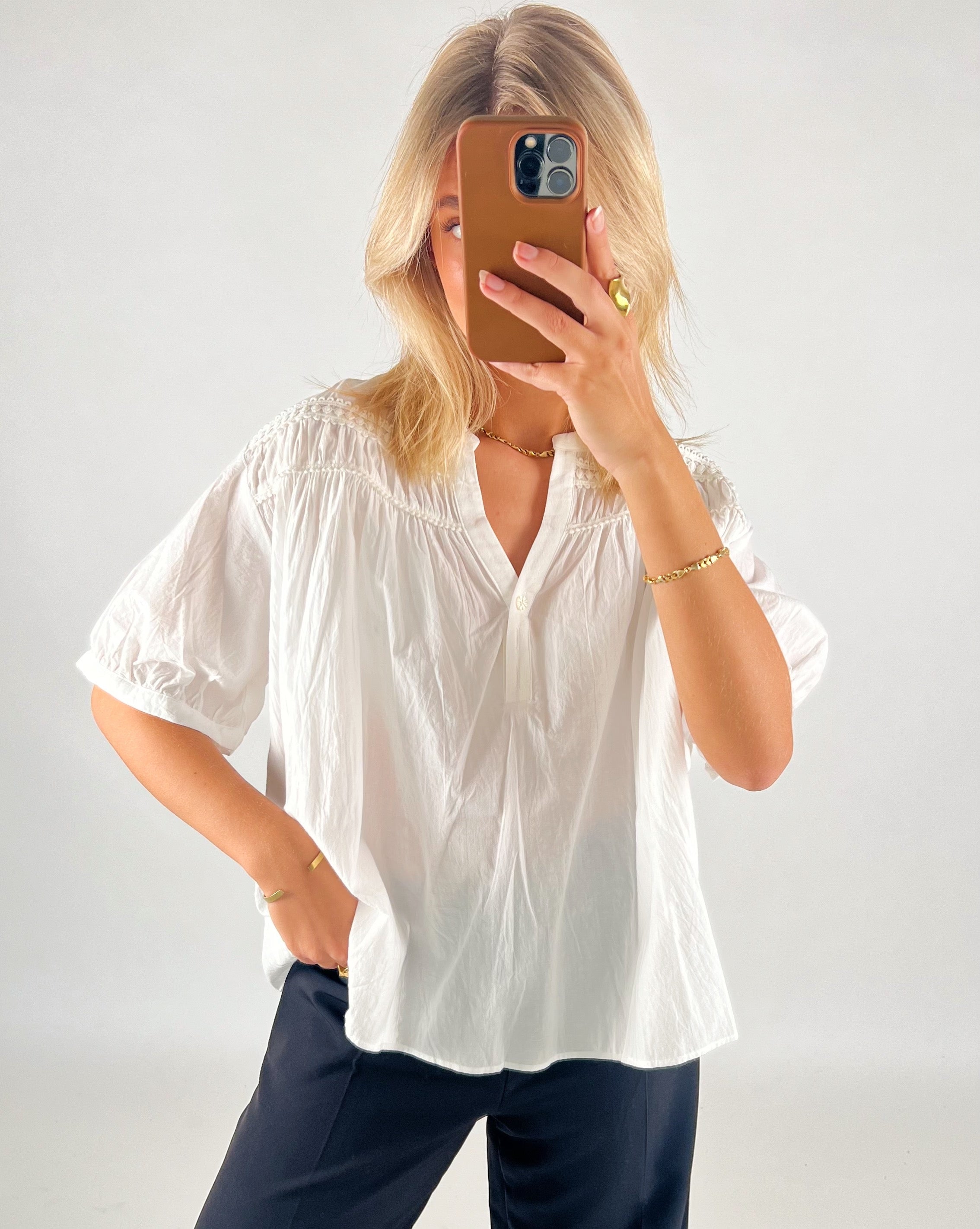 En person står i en hvid skjorte og sorte bukser, tager et spejlbillede med en telefon i brun etui, i et oplyst rum med hvid baggrund.