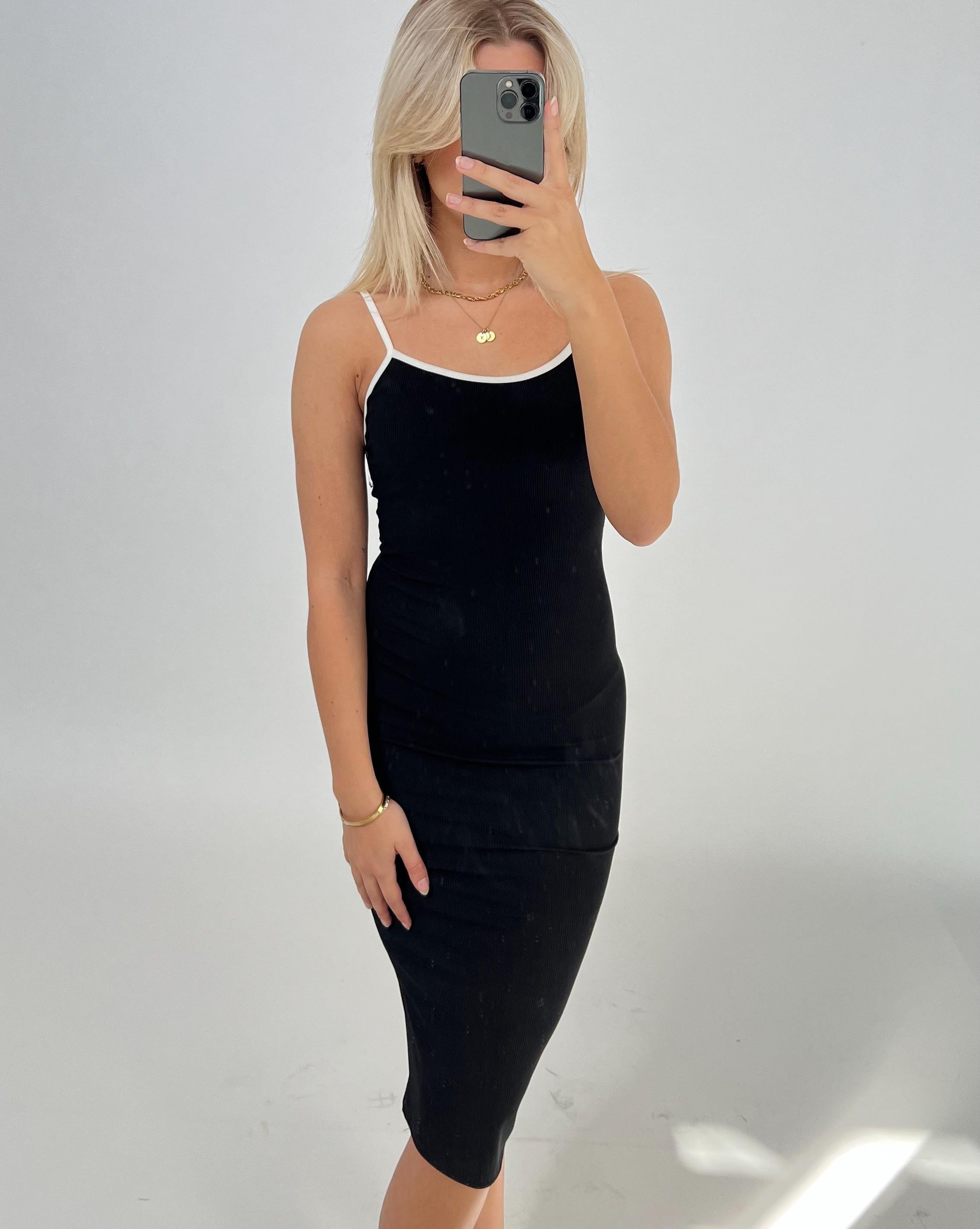 Person iført en sort kjole med hvide stropper tager et selfie i et spejl. Personen har lyst hår, og baggrunden er en enkel, lys væg.