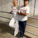 To personer i hvide T-shirts med teksten "GALAXY" står i en p-kælder. Den ene læner sig afslappet mod et gelænder, mens den anden har armene over kors.