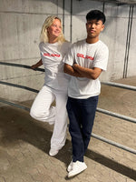 To personer i hvide T-shirts med teksten "GALAXY" står i en p-kælder. Den ene læner sig afslappet mod et gelænder, mens den anden har armene over kors.