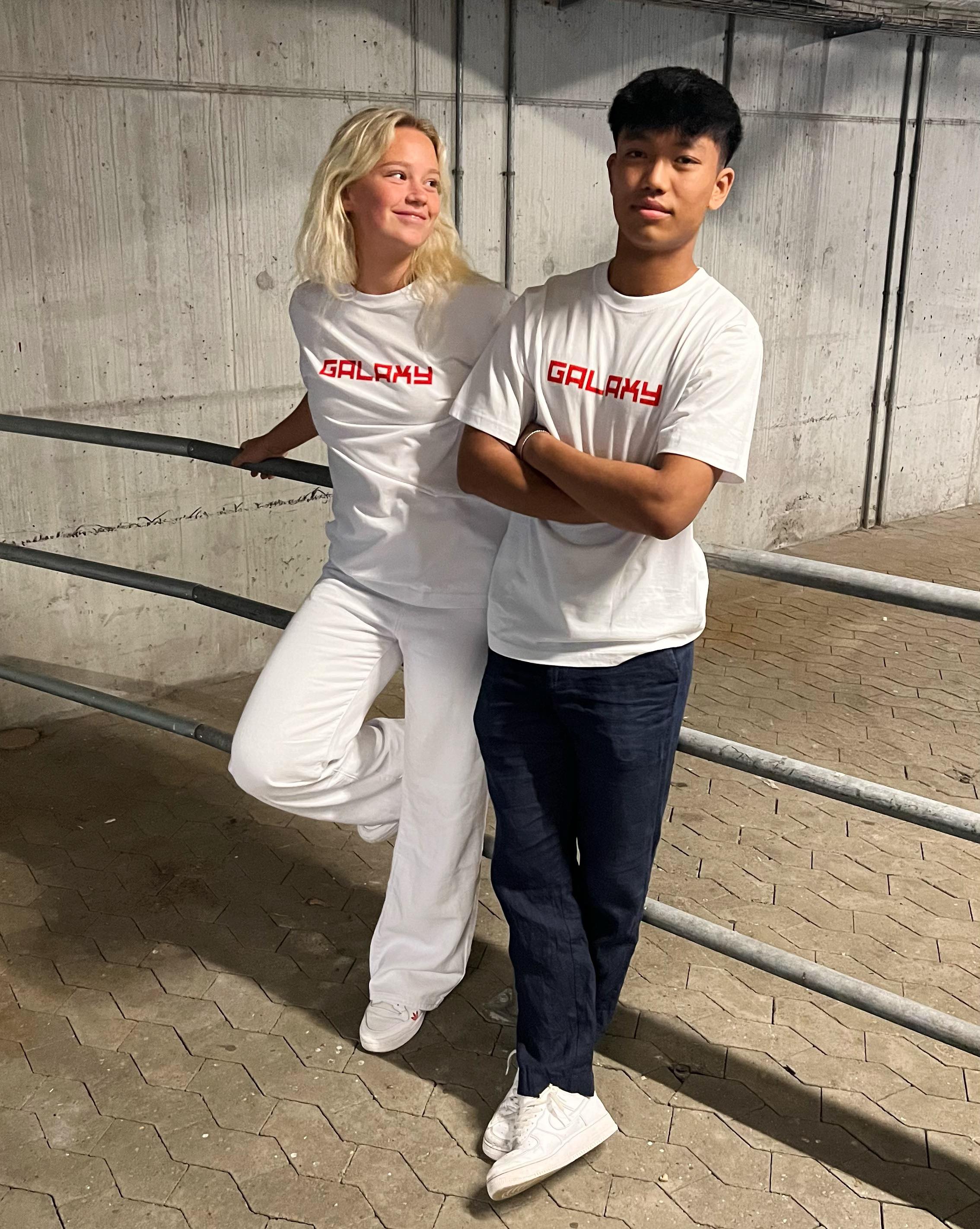 To personer i hvide T-shirts med teksten "GALAXY" står i en p-kælder. Den ene læner sig afslappet mod et gelænder, mens den anden har armene over kors.