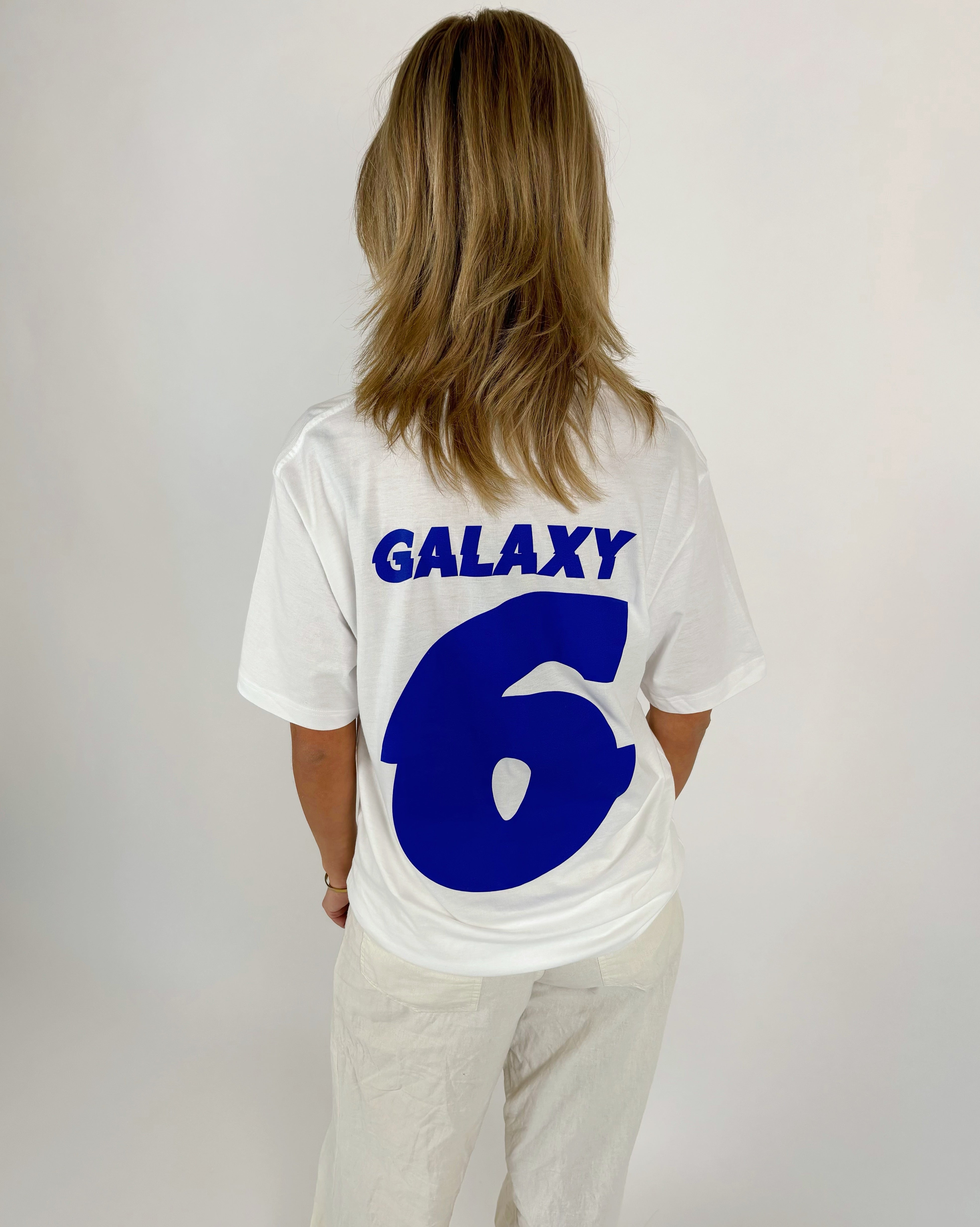 En person ses bagfra i en hvid T-shirt med blå tekst "GALAXY 6" i et lyst rum. Personen står stille med hænderne ved siden.