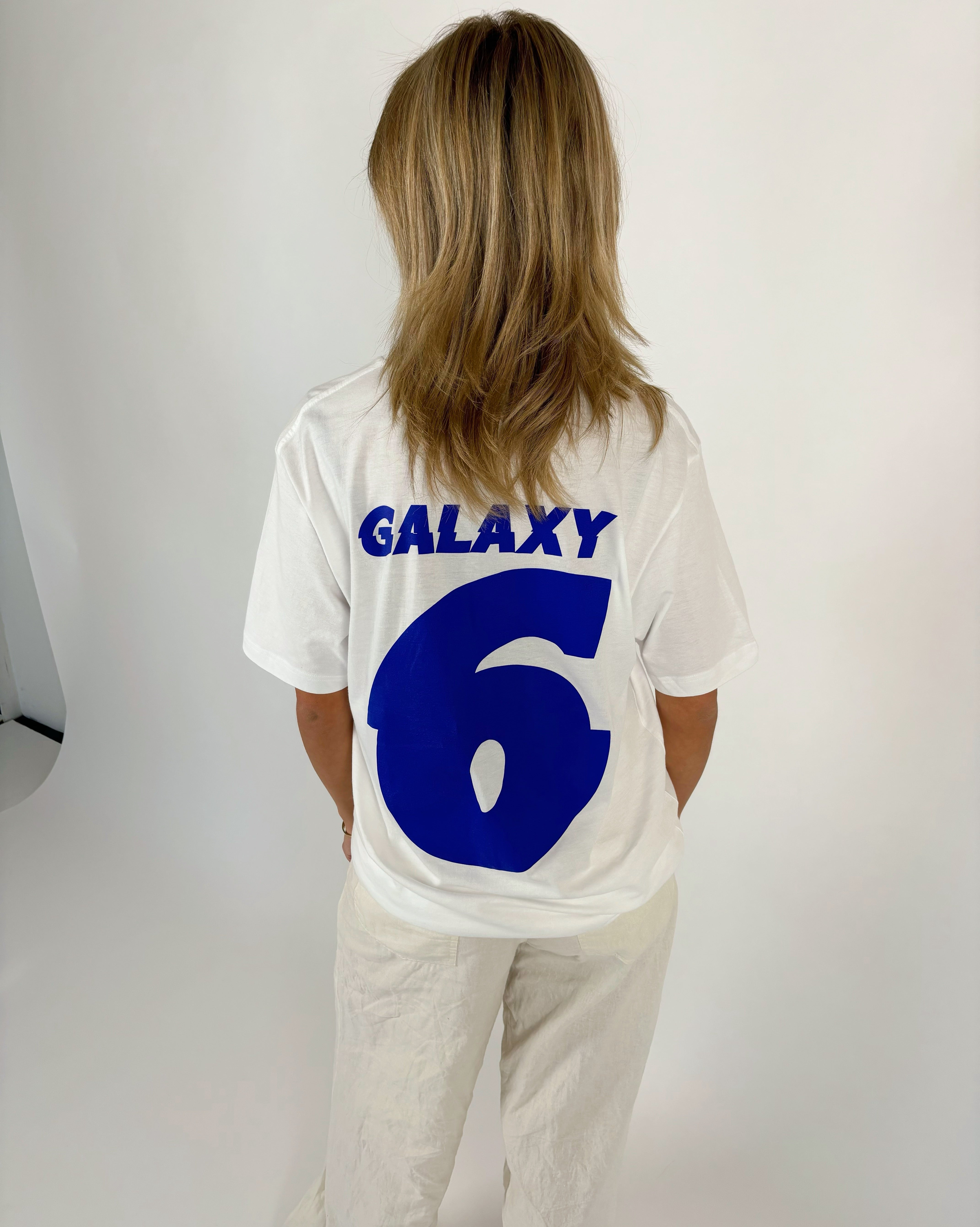 En person med skulderlangt hår står med ryggen til, iført en hvid t-shirt med blå tekst "GALAXY 6" i en lys baggrund.