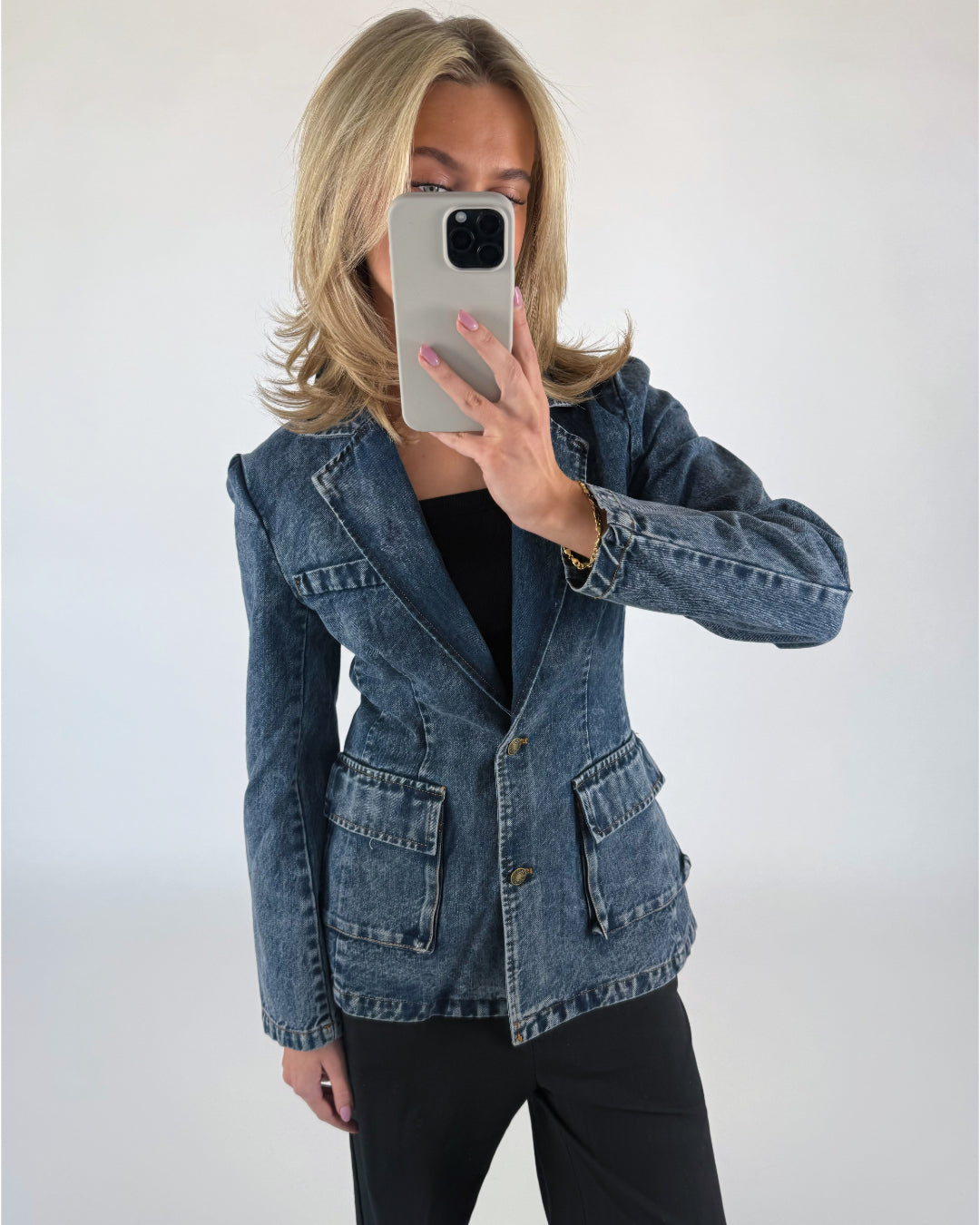 Person tager en spejlselfie iført en blå denimjakke med lommer, sorte bukser og har blondt hår. De holder en smartphone foran ansigtet i et neutralt baggrundsmiljø.