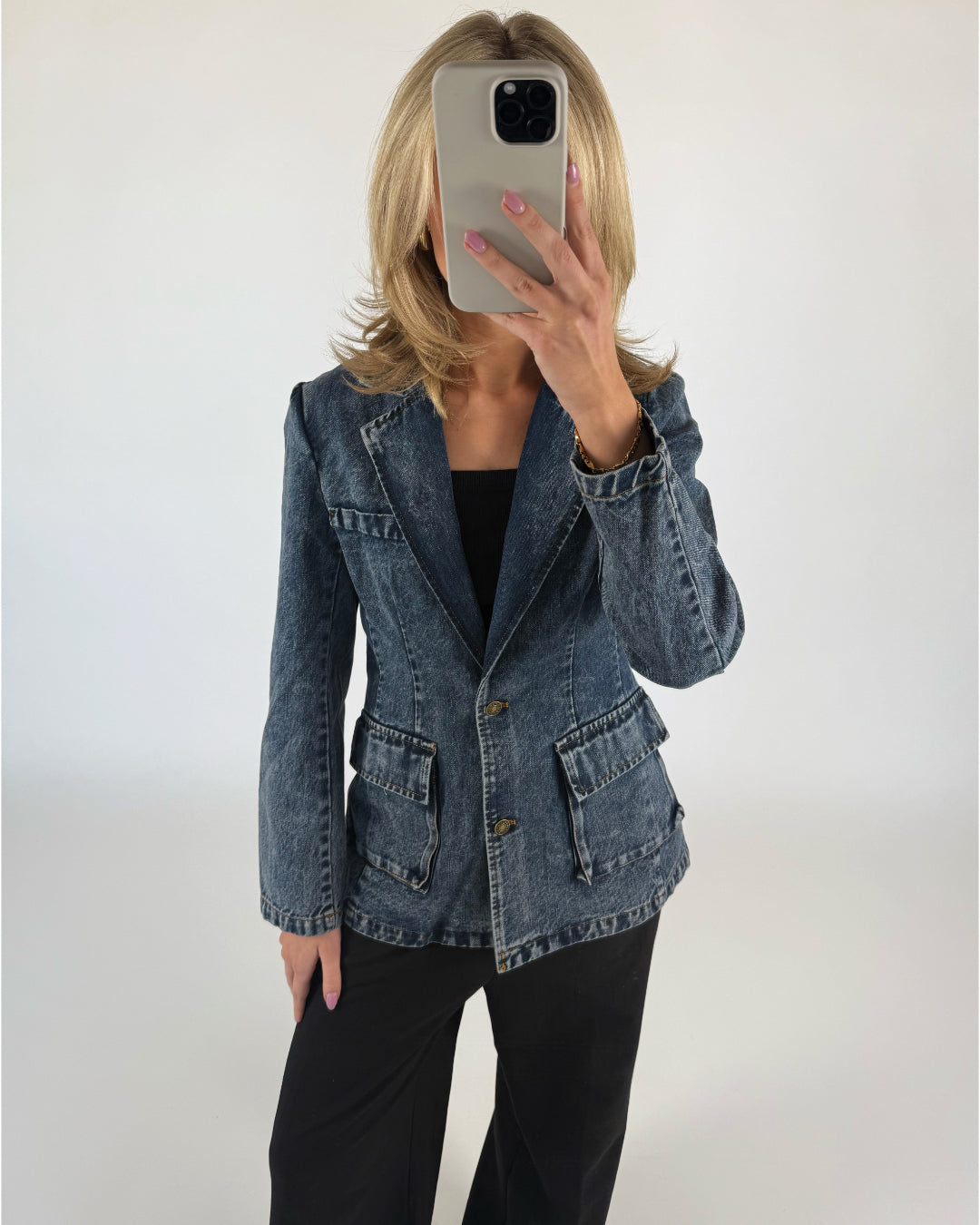 En person holder en smartphone foran ansigtet, iført en denimjakke med sorte bukser, står mod en hvid baggrund.
